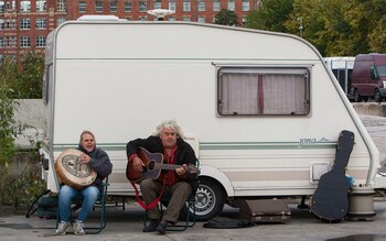 Two travellers play music on a guitar and tambourine outside their caravan